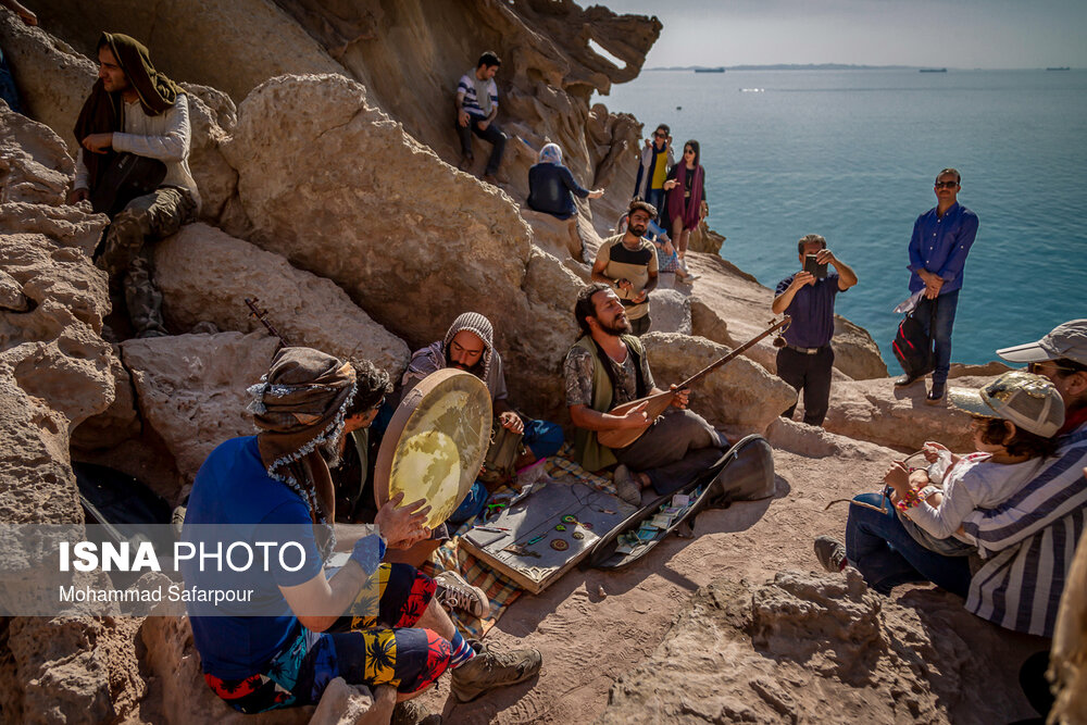 چند گردشگر نوازنده در حال نواختن ساز در دره مجسمه‌ها در جزیره هرمز هستند و مسافران در حال تماشا و تصویر برداری از آن‌ها هستند.